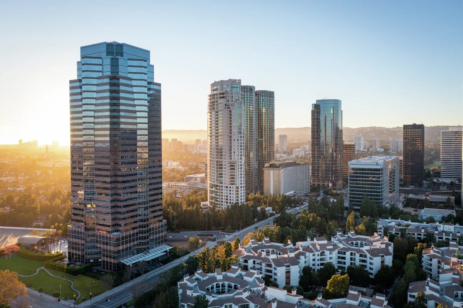 City skyline during sunset with tall modern skyscrapers, smaller residential buildings, and green trees in the foreground