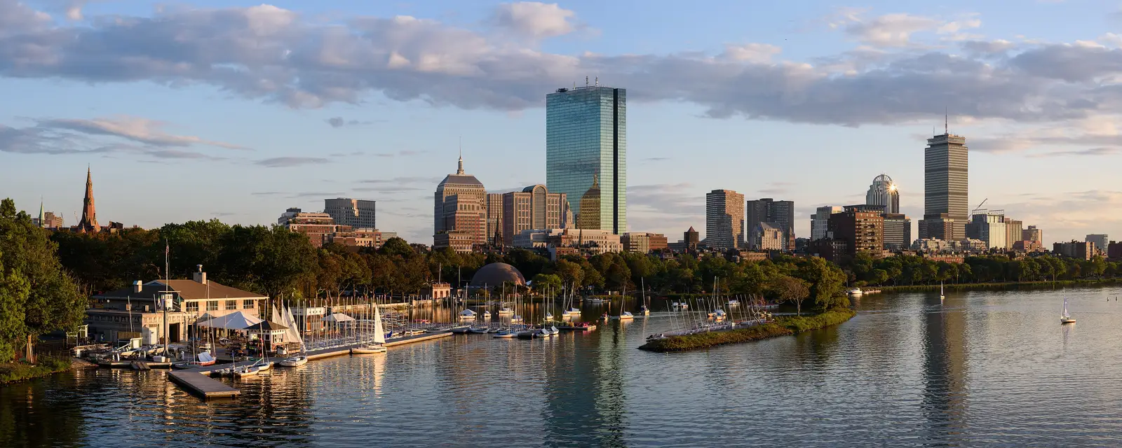 Panoramic golden-hour view of the Boston, Massachusetts skyline and Back Bay skyscrapers reflected in the Charles River, seen from the Longfellow Bridge